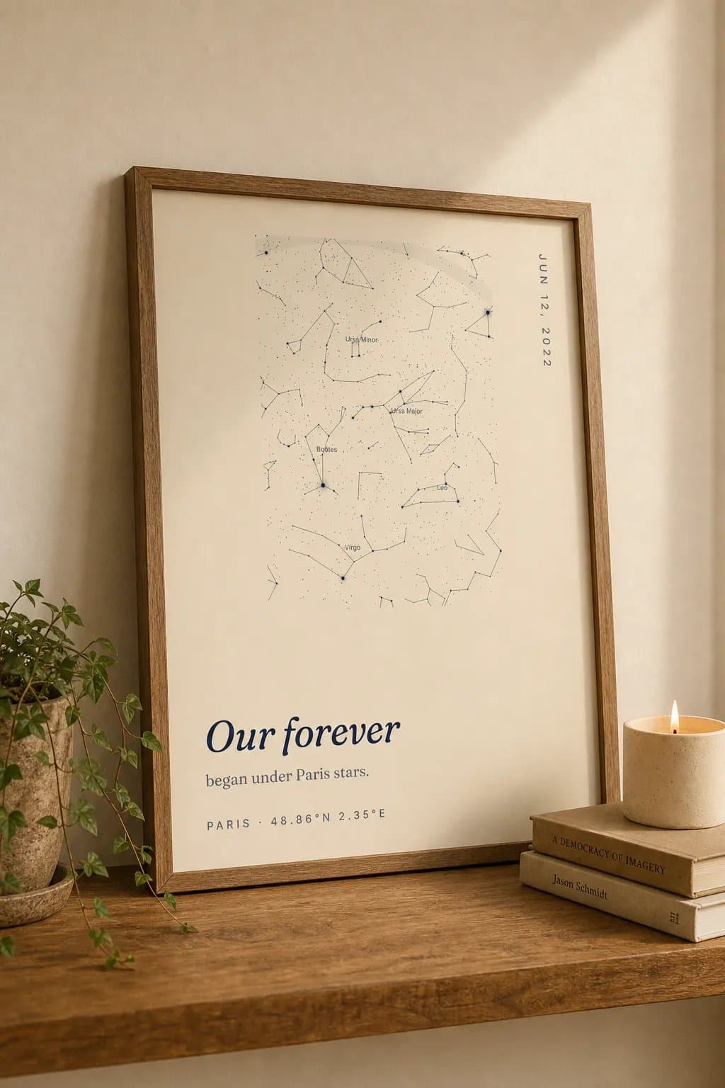 A framed cream star map titled Our forever leaning on a wooden shelf beside a candle and a small stack of books.
